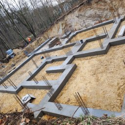 KLAMERBUD - Wylane betonowe fundamenty z wystającymi prętami zbrojeniowymi na piaszczystym podłożu, widok z góry, teren budowy na skraju lasu.