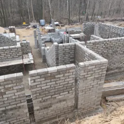 Wznoszone ściany fundamentowe z szarej cegły silikatowej na placu budowy, widoczne pręty zbrojeniowe, w tle las.