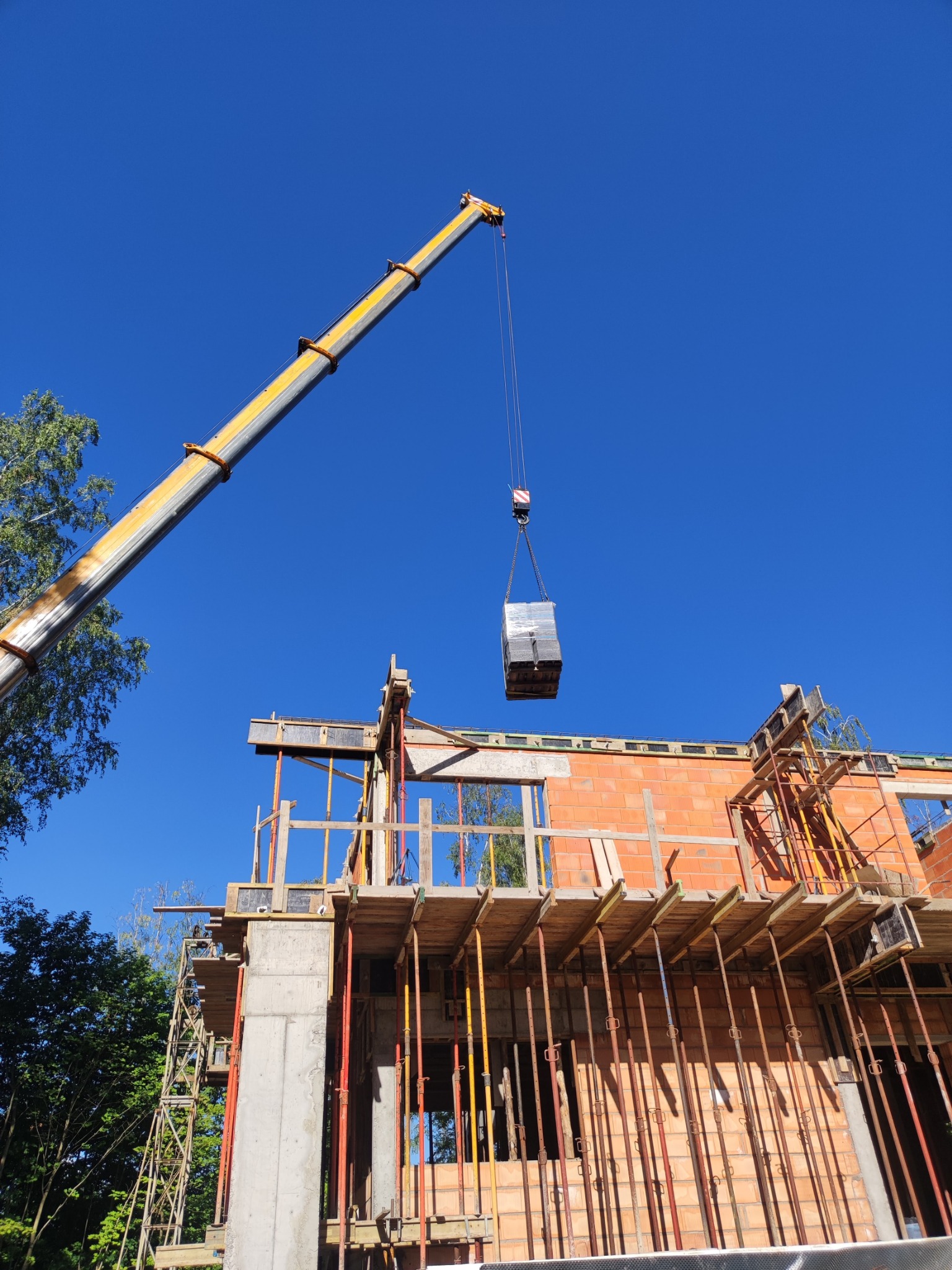 Żuraw budowlany podnosi pakiet materiałów na placu budowy domu w trakcie wznoszenia ścian, widoczne rusztowania i podpory, błękitne niebo w tle.