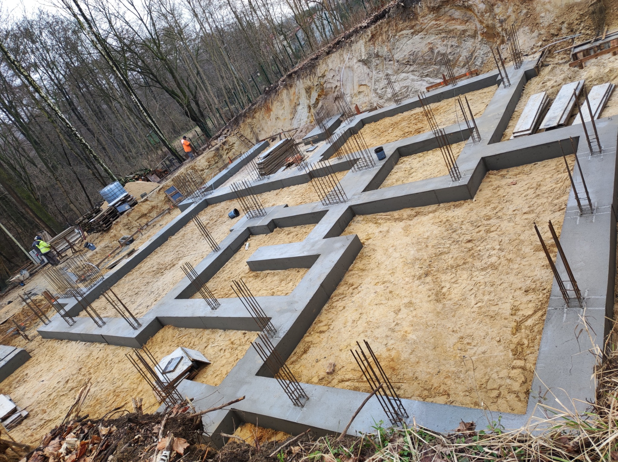 Wylane betonowe fundamenty z wystającymi prętami zbrojeniowymi na piaszczystym podłożu, widok z góry, teren budowy na skraju lasu.