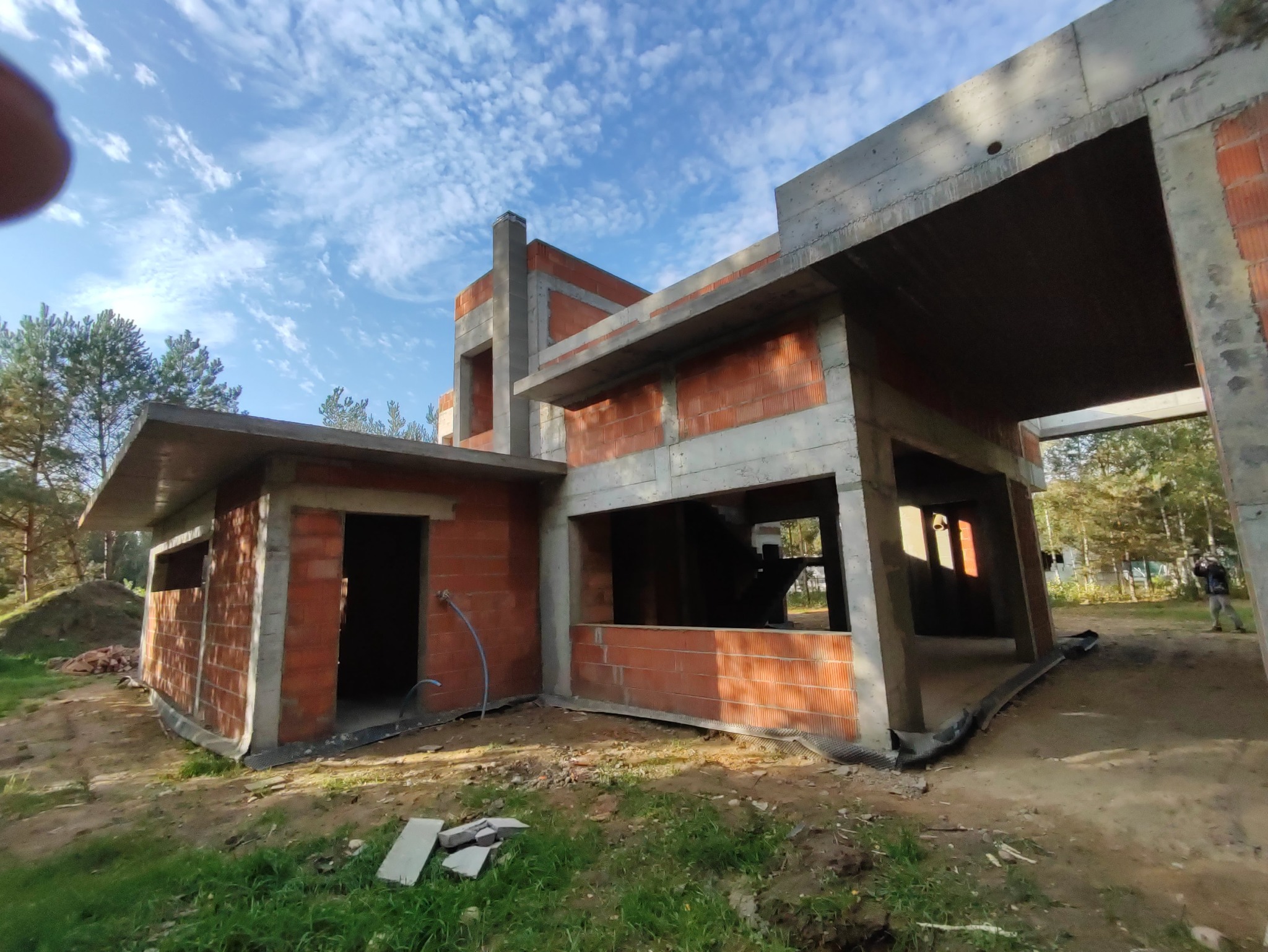 Surowy stan budynku z czerwonej cegły i betonu w lesistym otoczeniu, widoczne niezakończone ściany i betonowe nadproża, błękitne niebo w tle.