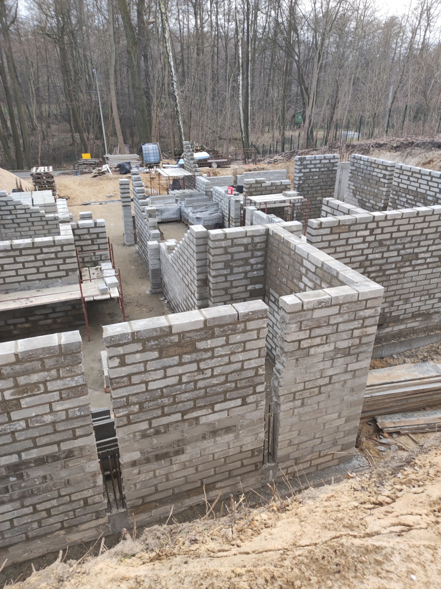 Wznoszone ściany fundamentowe z szarej cegły silikatowej na placu budowy, widoczne pręty zbrojeniowe, w tle las.