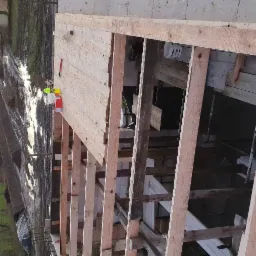 Wewnętrzny szkielet konstrukcji domu w trakcie budowy, widoczne drewniane belki, fragmenty ścian z betonu kom&oacute;rkowego i narzędzia malarskie na g&oacute;rnej krawędzi ściany.