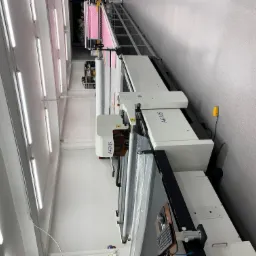 Automatyczna maszyna do cięcia materiałów tekstylnych w jasnym pomieszczeniu produkcyjnym, z rolką materiału i panelem sterowania, widoczne oświetlenie sufitowe.