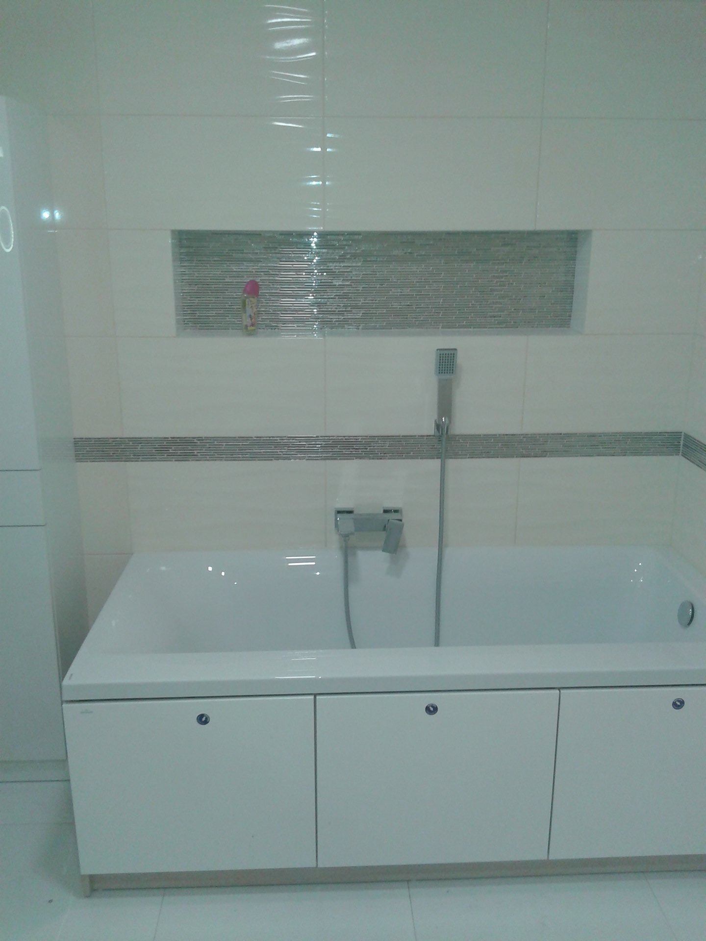 Biała wanna obudowana szafkami z chromowanymi uchwytami na tle wykafelkowanej ściany z wnęką dekoracyjną i szklaną mozaiką.