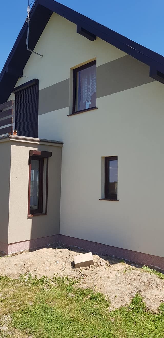 Jasna elewacja domu z brązowymi oknami i roletami, widoczny fragment dachu, trawnik i piasek przy ścianie, element dekoracyjny w oknie.