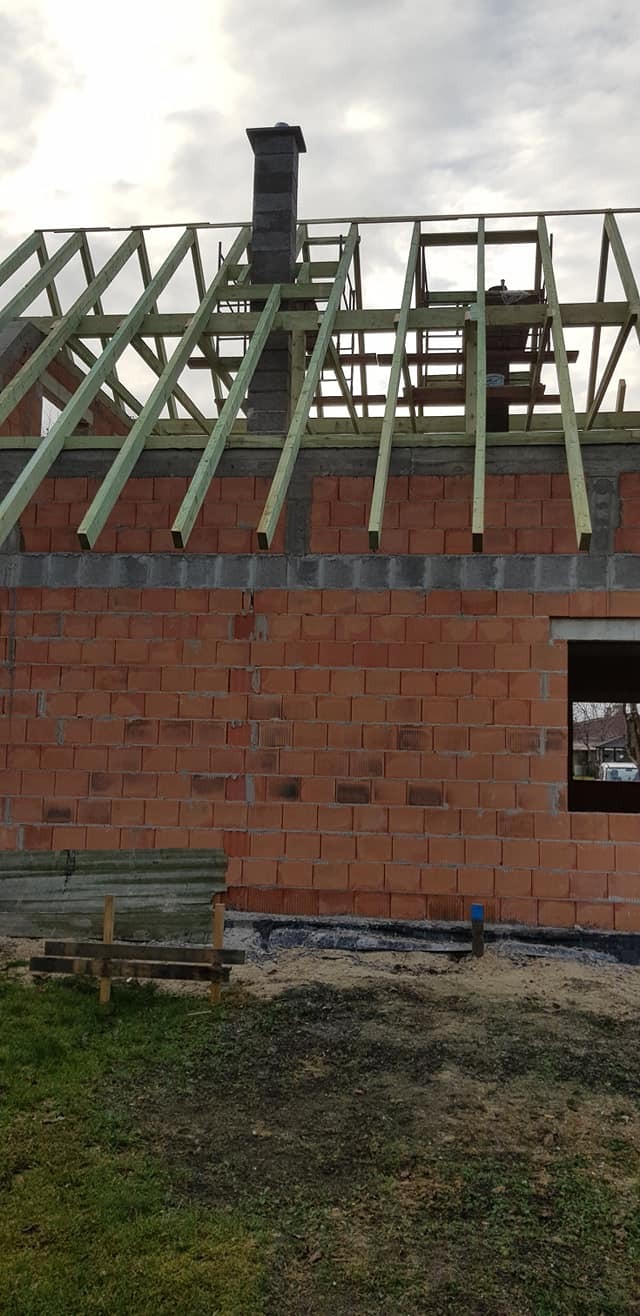 Wznoszenie domu z czerwonej cegły: widoczna murowana ściana, drewniana konstrukcja dachu w trakcie montażu oraz komin z szarej cegły, teren wokół w stanie surowym.