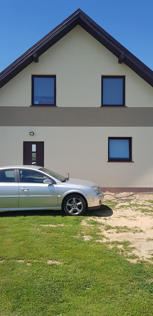 Dwukondygnacyjny dom z jasną elewacją, ciemnymi oknami i dachem, z boku zaparkowany srebrny samochód osobowy, widoczny fragment trawnika.