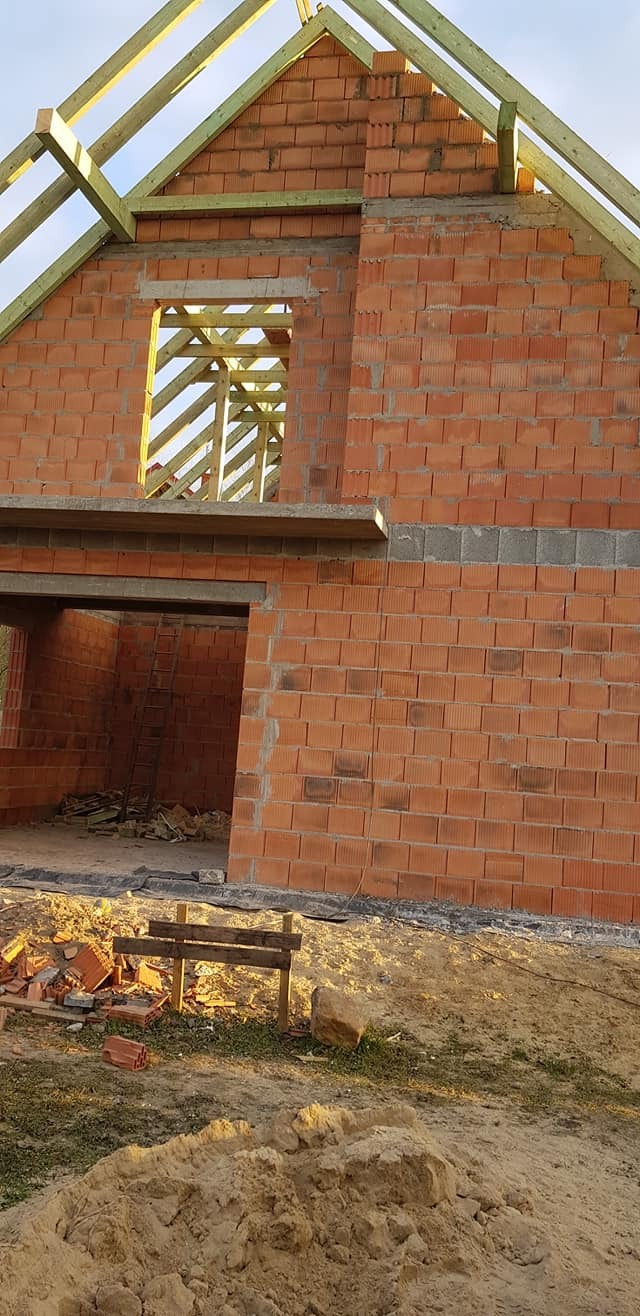 Surowy stan domu z czerwonej cegły, widoczne drewniane krokwie dachu i otwory okienne, zwały piasku i resztki cegieł na placu budowy.