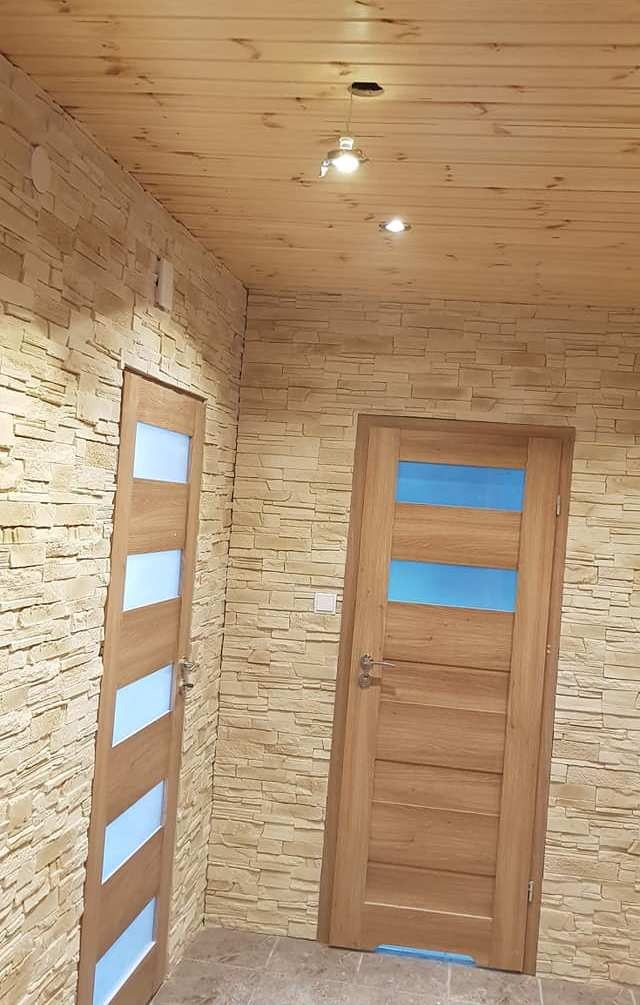 Wnętrze z dwoma drewnianymi drzwiami z niebieskimi szybami, ściany wykończone imitacją kamienia, drewniany sufit z oświetleniem punktowym.