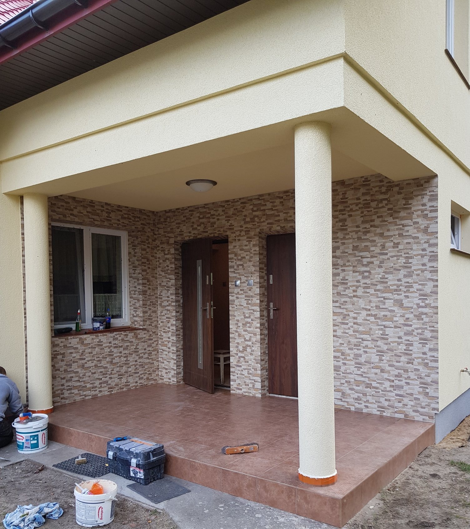 Wykończone wejście do domu z elewacją w odcieniach beżu, brązowe drzwi i płytki na podłodze werandy, widoczne narzędzia i wiaderka z farbą sugerują prace wykończeniowe.