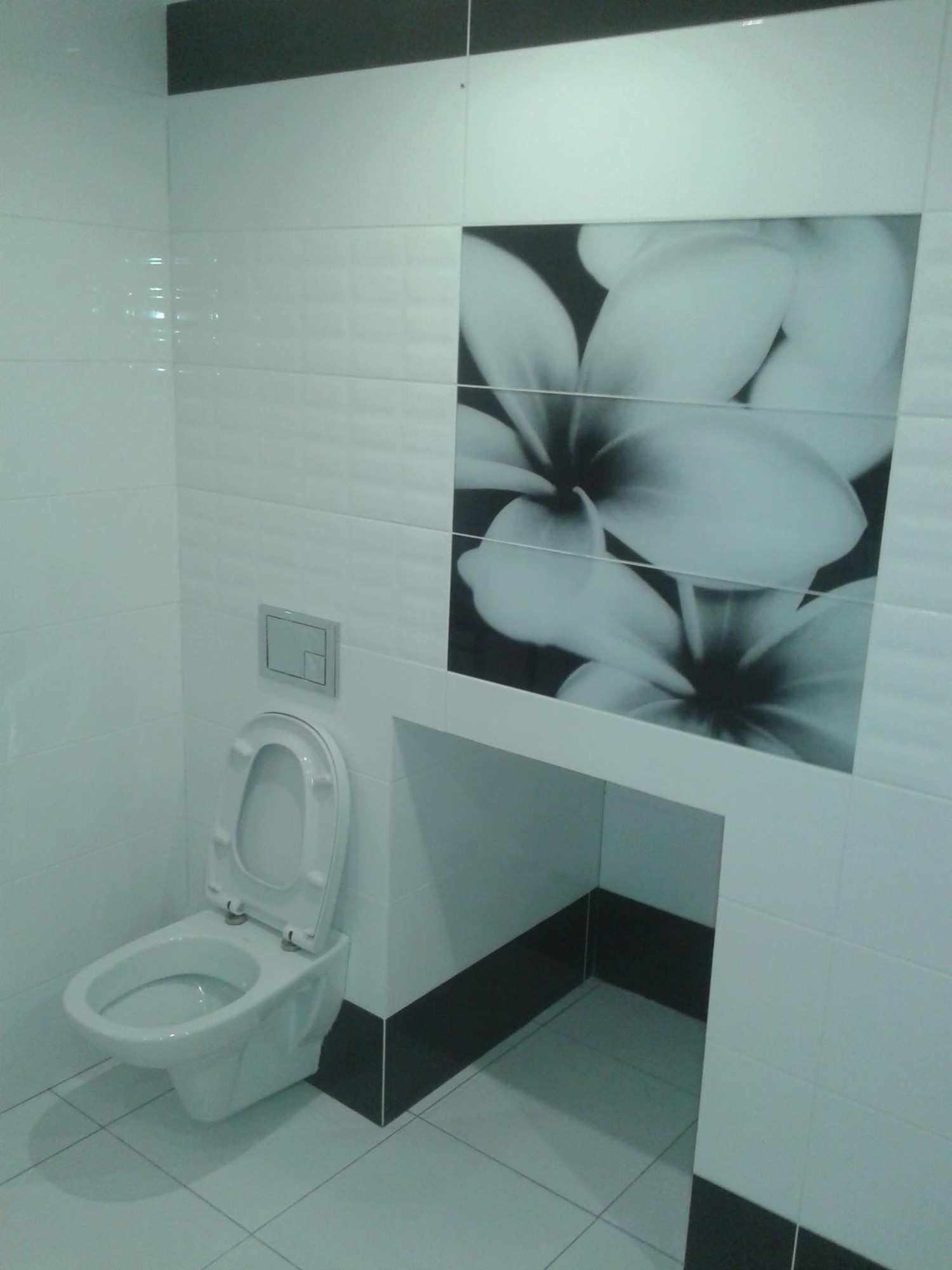 Biała wisząca toaleta z otwartą deską w nowoczesnej łazience wyłożonej białymi płytkami z dekoracyjnym panelem z motywem kwiatowym w czerni i bieli.