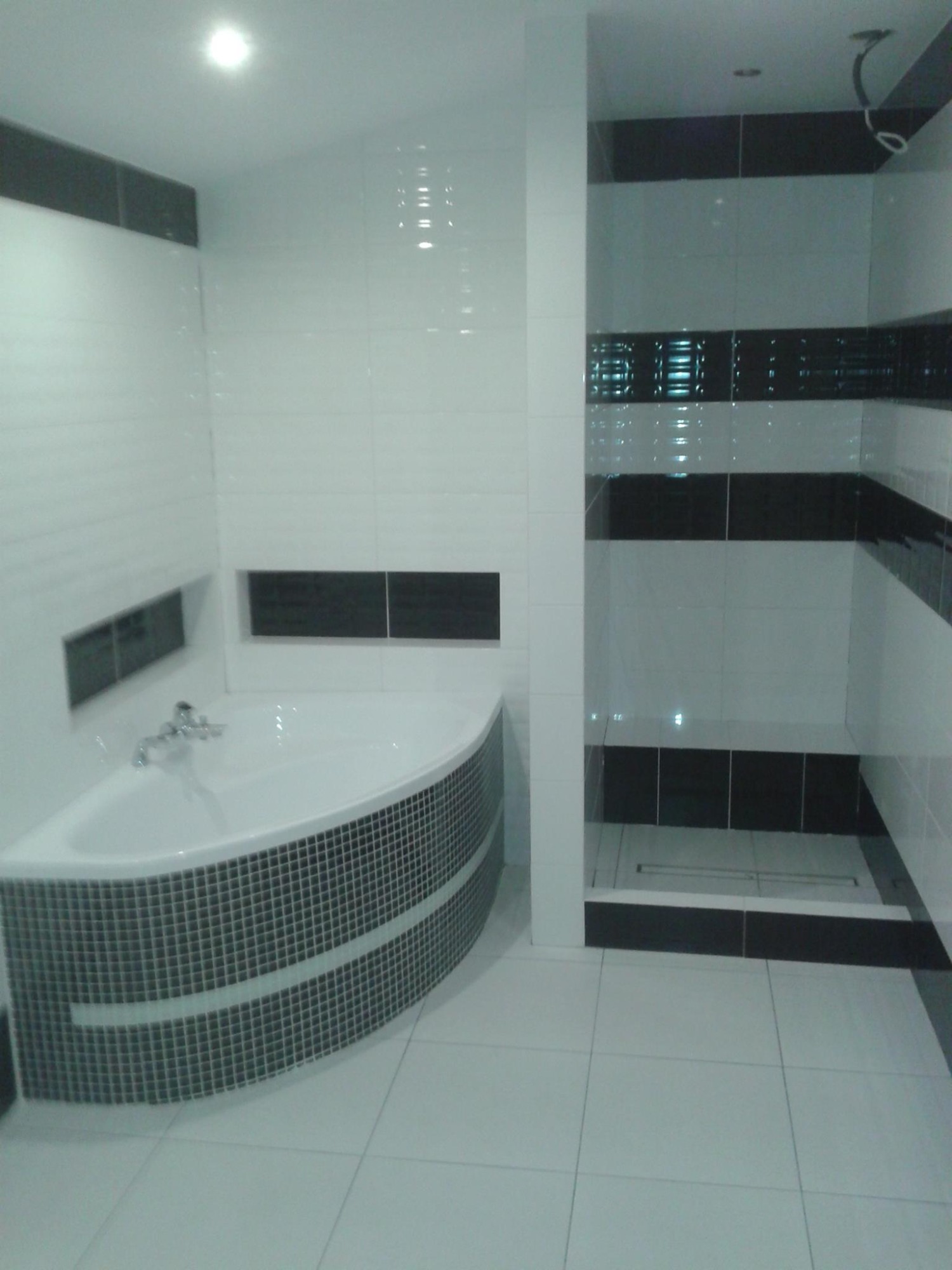 Nowoczesna łazienka z biało-czarnymi płytkami, narożna wanna obudowana mozaiką, kabina prysznicowa typu walk-in z odpływem liniowym.