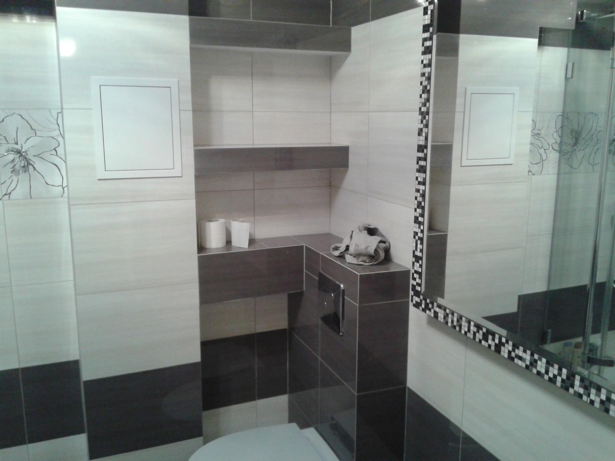 Nowoczesna toaleta z podtynkową spłuczką, wykończona płytkami w odcieniach bieli i szarości z dekoracyjnym motywem kwiatowym, lustro z mozaikową ramą.