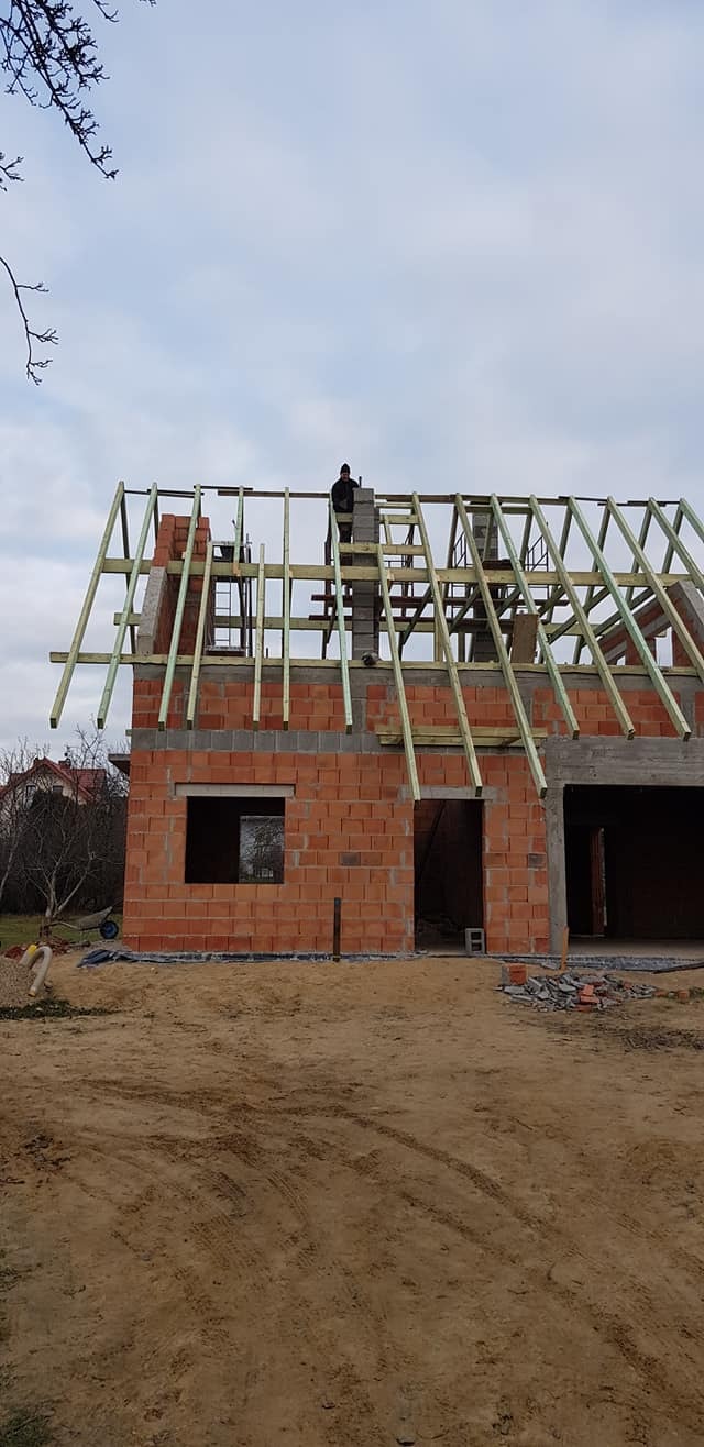 Surowy stan domu z czerwonej cegły w trakcie budowy, widoczny szkielet dachu i murarz pracujący nad kominem, ziemisty teren wokół.