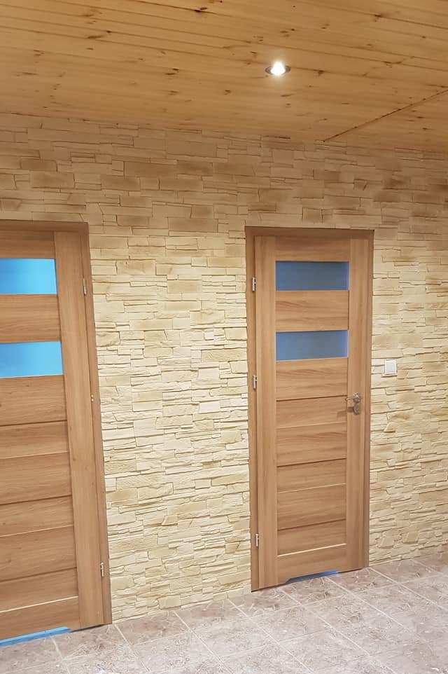 Dwa drewniane drzwi z przeszkleniami w odcieniach niebieskiego, zamontowane w ścianie wykończonej dekoracyjnym kamieniem imitującym piaskowiec, pod drewnianym sufitem.
