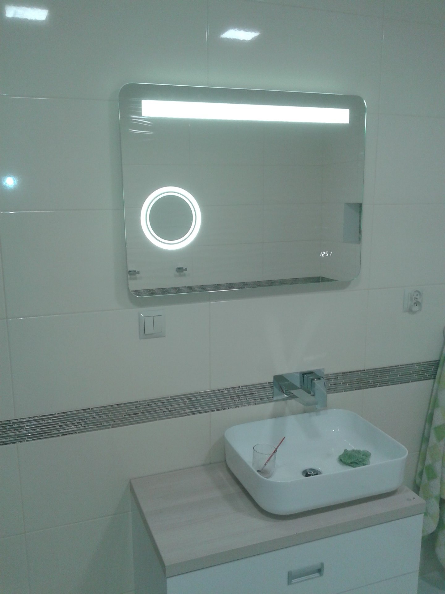 Nowoczesna łazienka z umywalką nablatową i podświetlanym lustrem z wyświetlaczem czasu, wykończona jasnymi płytkami z dekoracyjnym pasem.