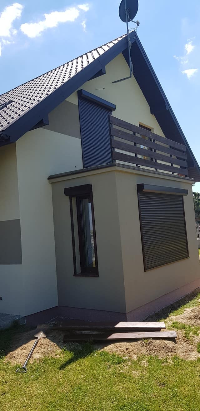 Dwukondygnacyjny dom z beżową elewacją i brązowymi roletami zewnętrznymi, widoczne elementy drewniane balkonu i brązowa dachówka, antena satelitarna na dachu, na trawniku leży łopata.