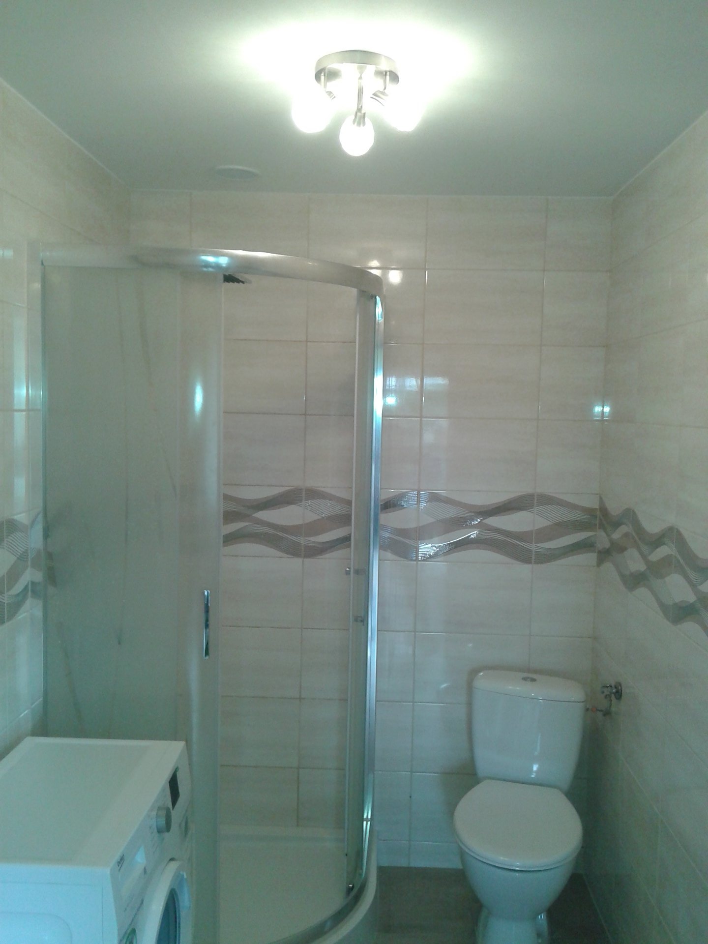 Wykończona łazienka z kabiną prysznicową narożną, pralką i toaletą, wyłożona jasnymi płytkami ceramicznymi z dekoracyjnym pasem w kształcie fal.