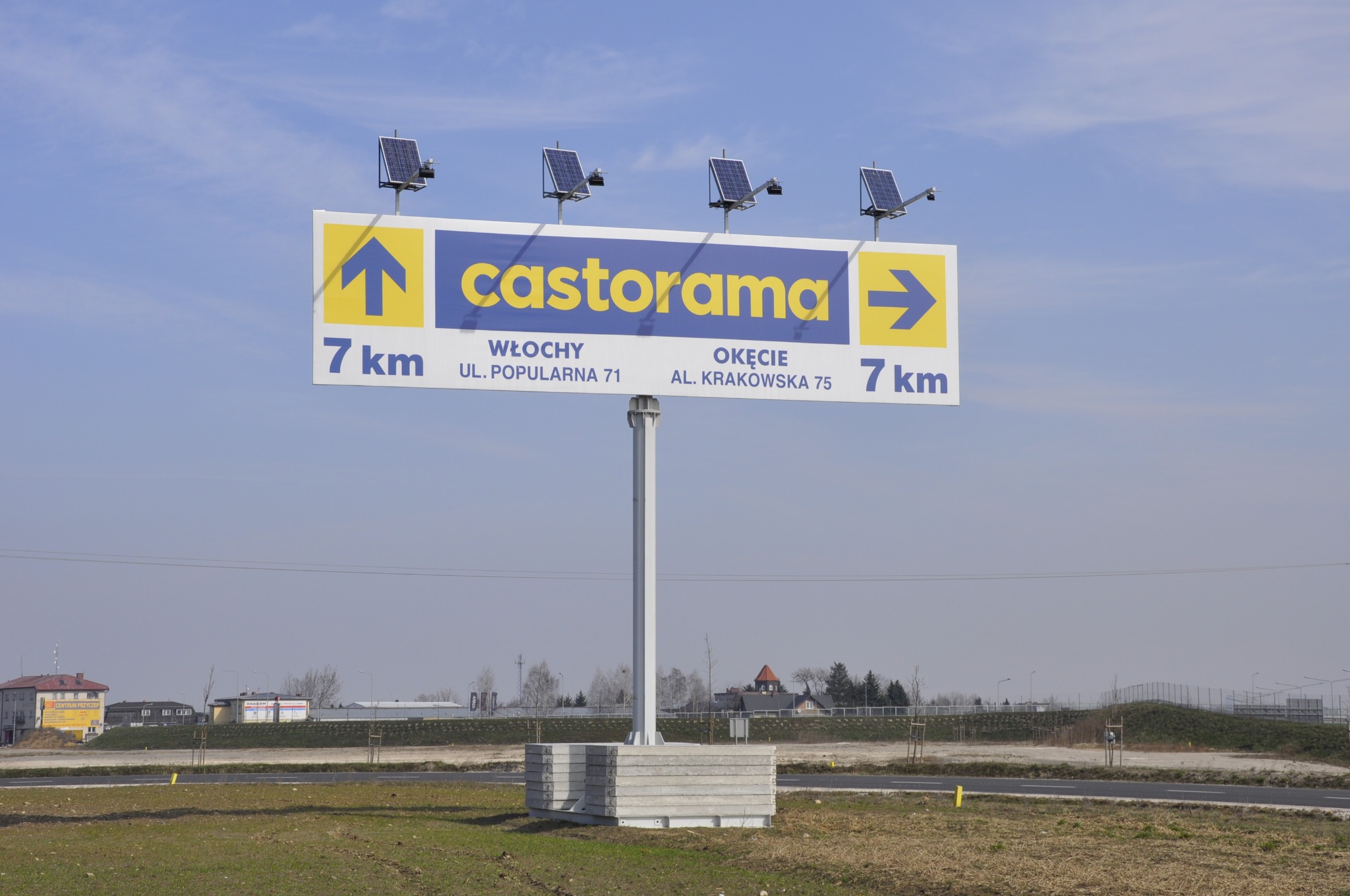 Billboard z logo Castorama, wskazujący odległość 7 km do Włoch i Okęcia, zasilany panelami słonecznymi, umieszczony na tle błękitnego nieba i krajobrazu.