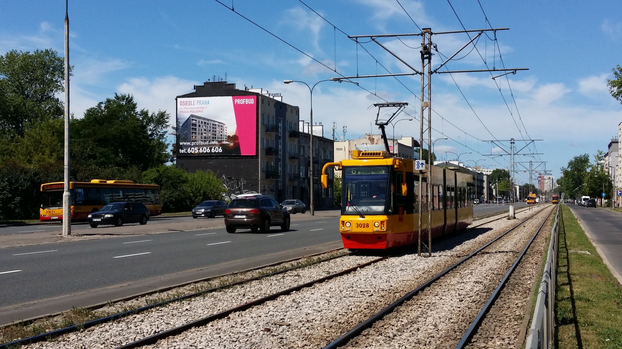 Żółto-czerwony tramwaj na torach w miejskiej scenerii, z billboardem reklamowym osiedla w tle.