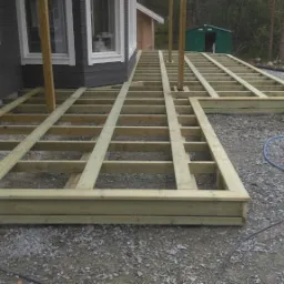 Szkielet konstrukcji drewnianej tarasu w trakcie budowy, widoczne drewniane legary i słupy, na żwirowym podłożu, w tle dom z szarą elewacją i białą stolarką okienną i drzwiową, narzędzia budowlane...