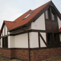 Dwukondygnacyjny dom w stylu muru pruskiego z czerwonym dachem, widoczne ceglane fundamenty i fragment rynny.