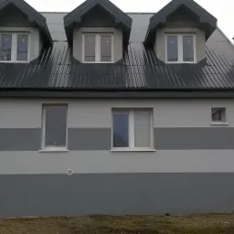 Odnowiona fasada budynku z szarą, poziomą pasową dekoracją, nowa blacha na dachu, trzy okna dachowe z widocznymi odbiciami drzew.