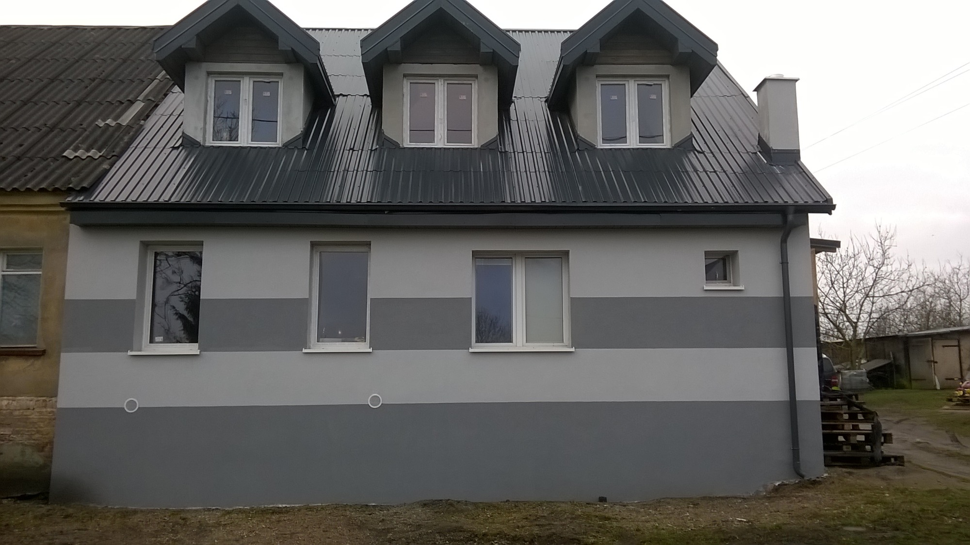 Odnowiona fasada budynku z szarą, poziomą pasową dekoracją, nowa blacha na dachu, trzy okna dachowe z widocznymi odbiciami drzew.