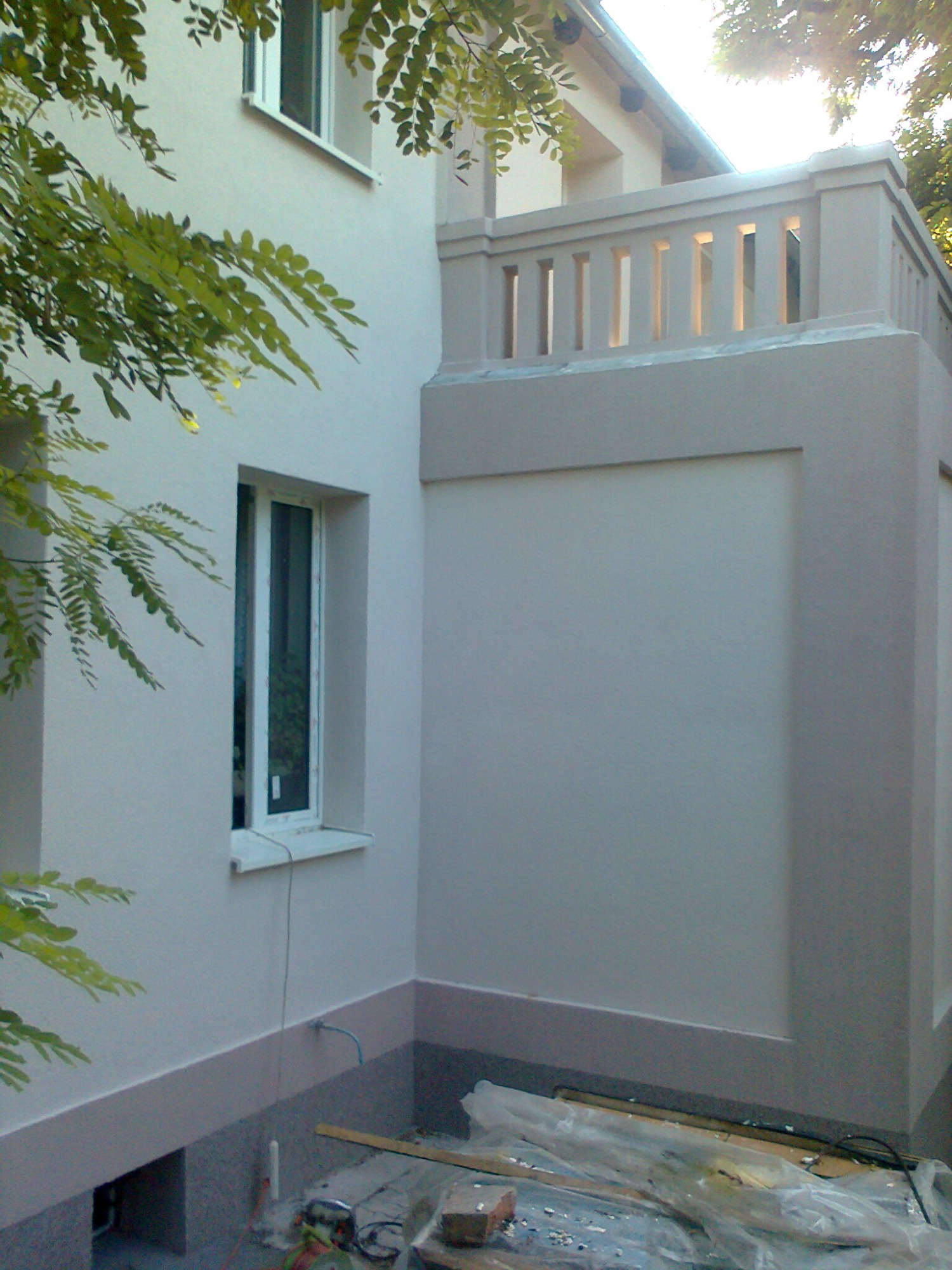 Świeżo otynkowana elewacja budynku z balkonem, widoczne okna i narzędzia budowlane w dolnej części zdjęcia.