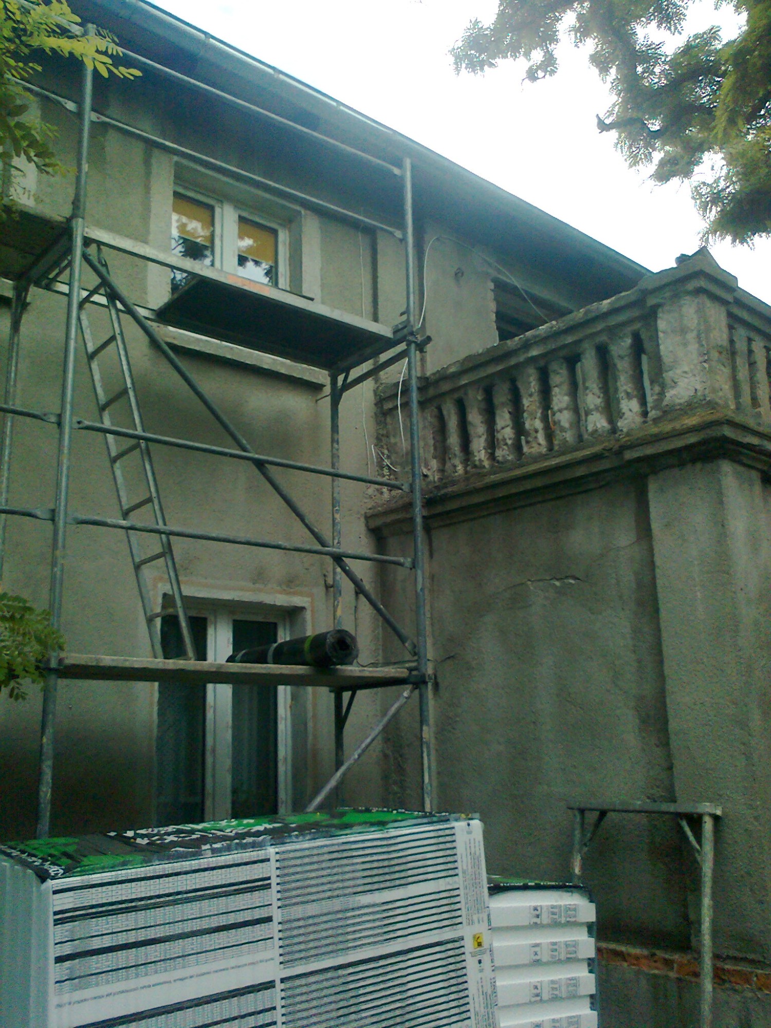 Szare rusztowanie przy szarej, starej elewacji budynku z balkonem; na rusztowaniu rolka papy, a u dołu paczki styropianu.