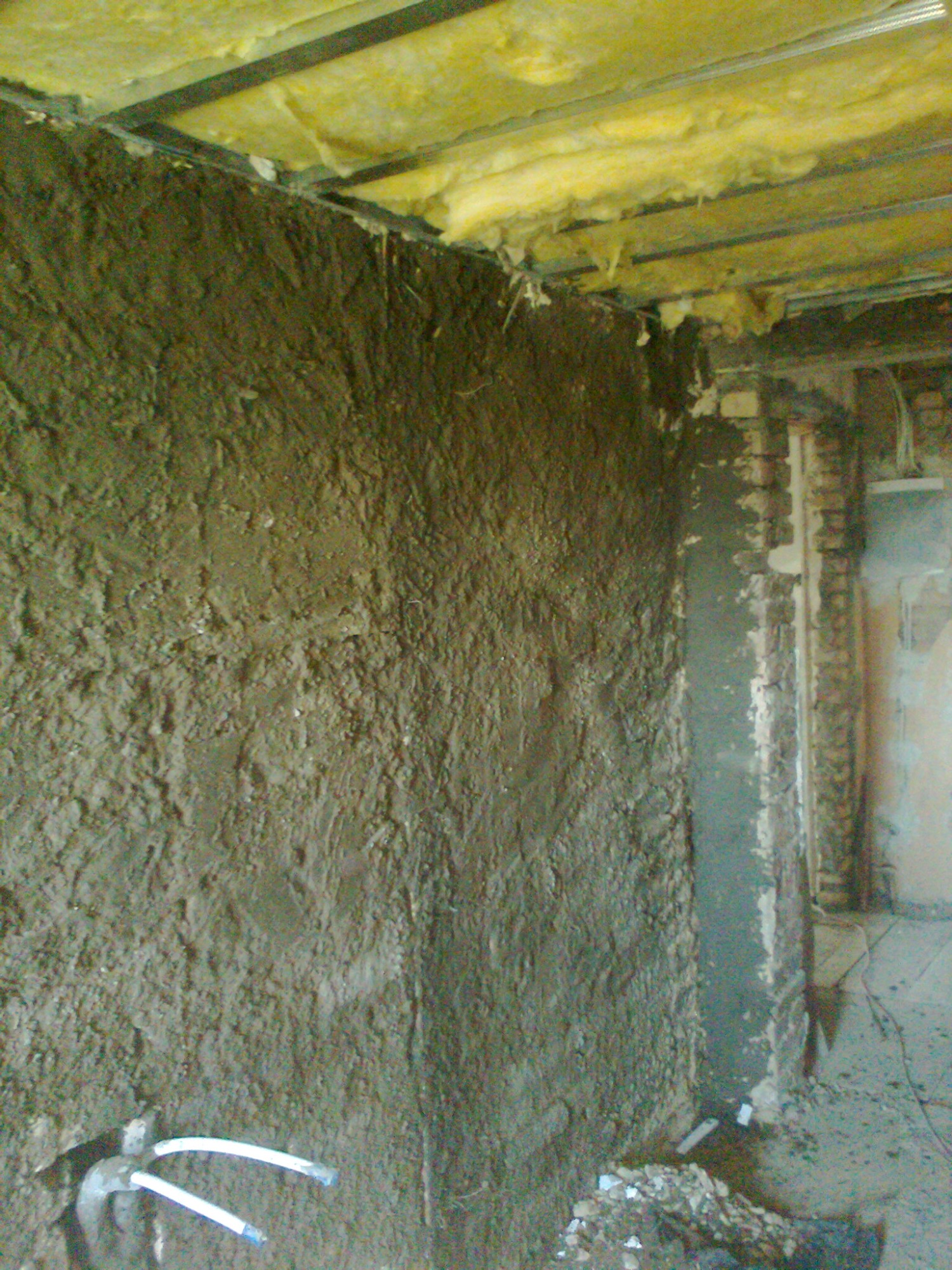 Surowa, nierówna ściana pokryta grubą warstwą świeżo nałożonej zaprawy, widoczne rury instalacji i fragment izolacji sufitu w trakcie prac remontowych.