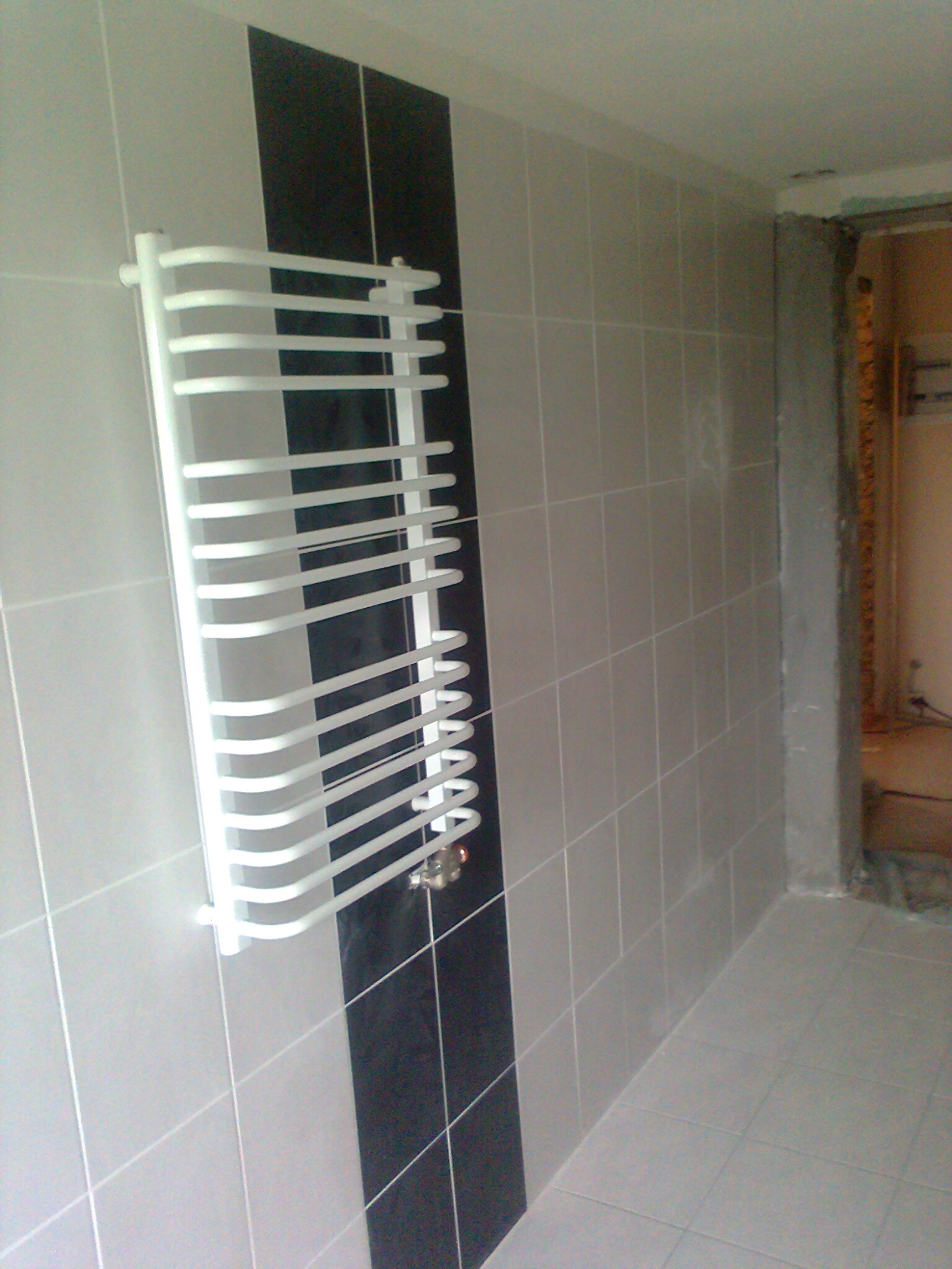 Biały grzejnik drabinkowy zamontowany na ścianie z szarych i czarnych płytek w łazience w trakcie remontu, widoczny fragment wejścia do pomieszczenia.