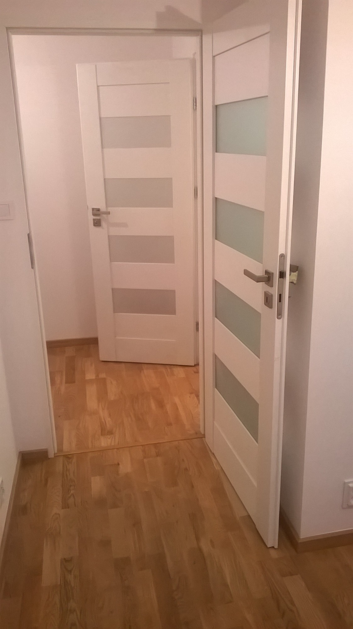 Dwa białe, nowoczesne drzwi wewnętrzne z przeszkleniami w poziomych pasach, zamontowane w jasnym pomieszczeniu z parkietem.