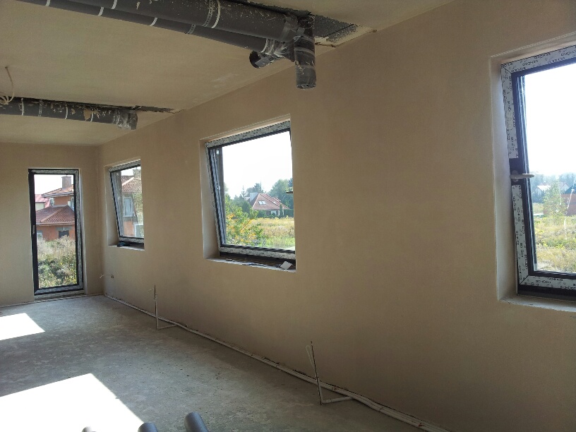 Wnętrze domu w stanie deweloperskim po nałożeniu gładzi gipsowej, widoczne okna z widokiem na zewnątrz, rury wentylacyjne pod sufitem oraz wystające przewody elektryczne przy podłodze.