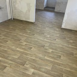 FLOORBUT SP. z o.o. - Jasna, drewnopodobna podłoga ułożona w nowym, przestronnym wnętrzu. Ściany częściowo przygotowane do malowania. Widok na kolejne pomieszczenie.