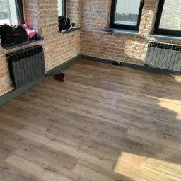 Nowa, drewnopodobna podłoga w pokoju z ceglanymi ścianami i grzejnikami pod oknami, widoczne buty na podłodze.