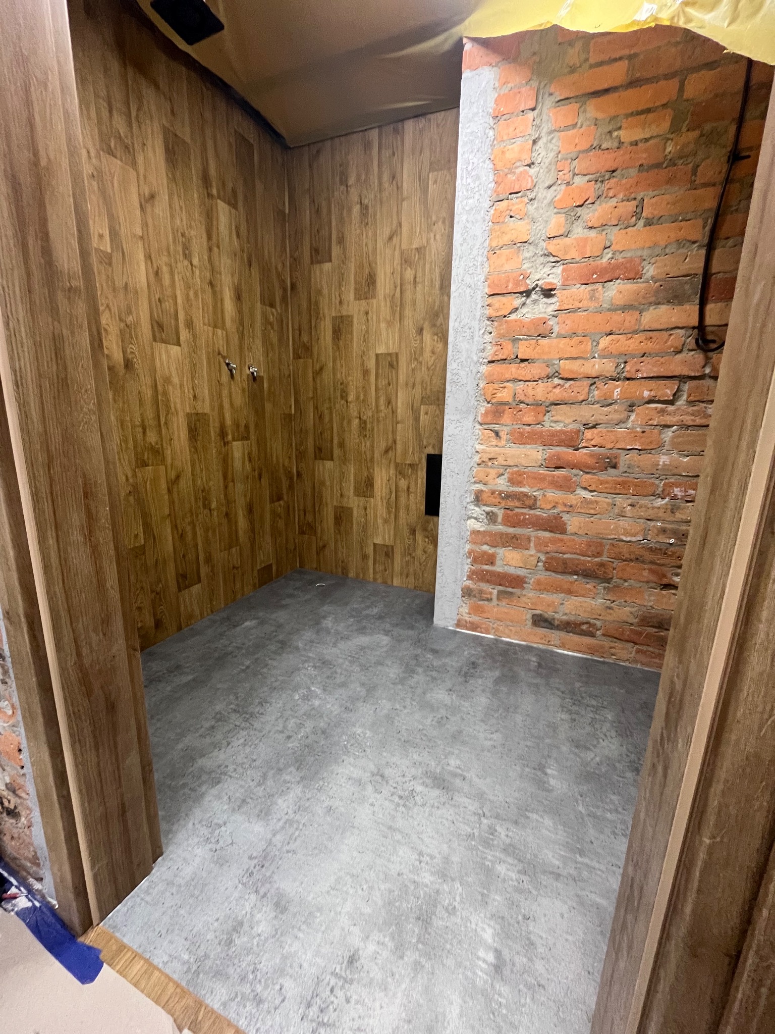 Wnętrze w trakcie remontu: drewnopodobne panele ścienne, surowa cegła, szara posadzka. Widoczne elementy instalacji i zabezpieczenia folią malarską.