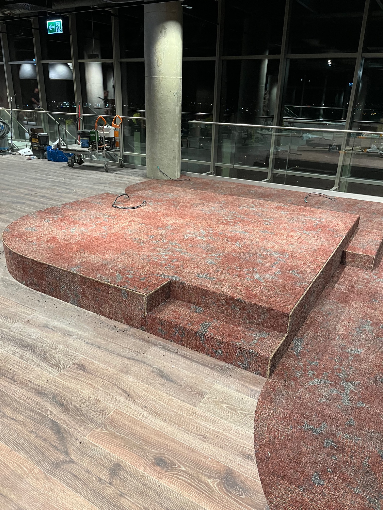 Wielopoziomowa, czerwona wykładzina dywanowa z wzorem, ułożona na drewnianej podłodze w nowoczesnym wnętrzu z dużymi oknami i kolumną. Wykładzina tworzy schody i platformę.