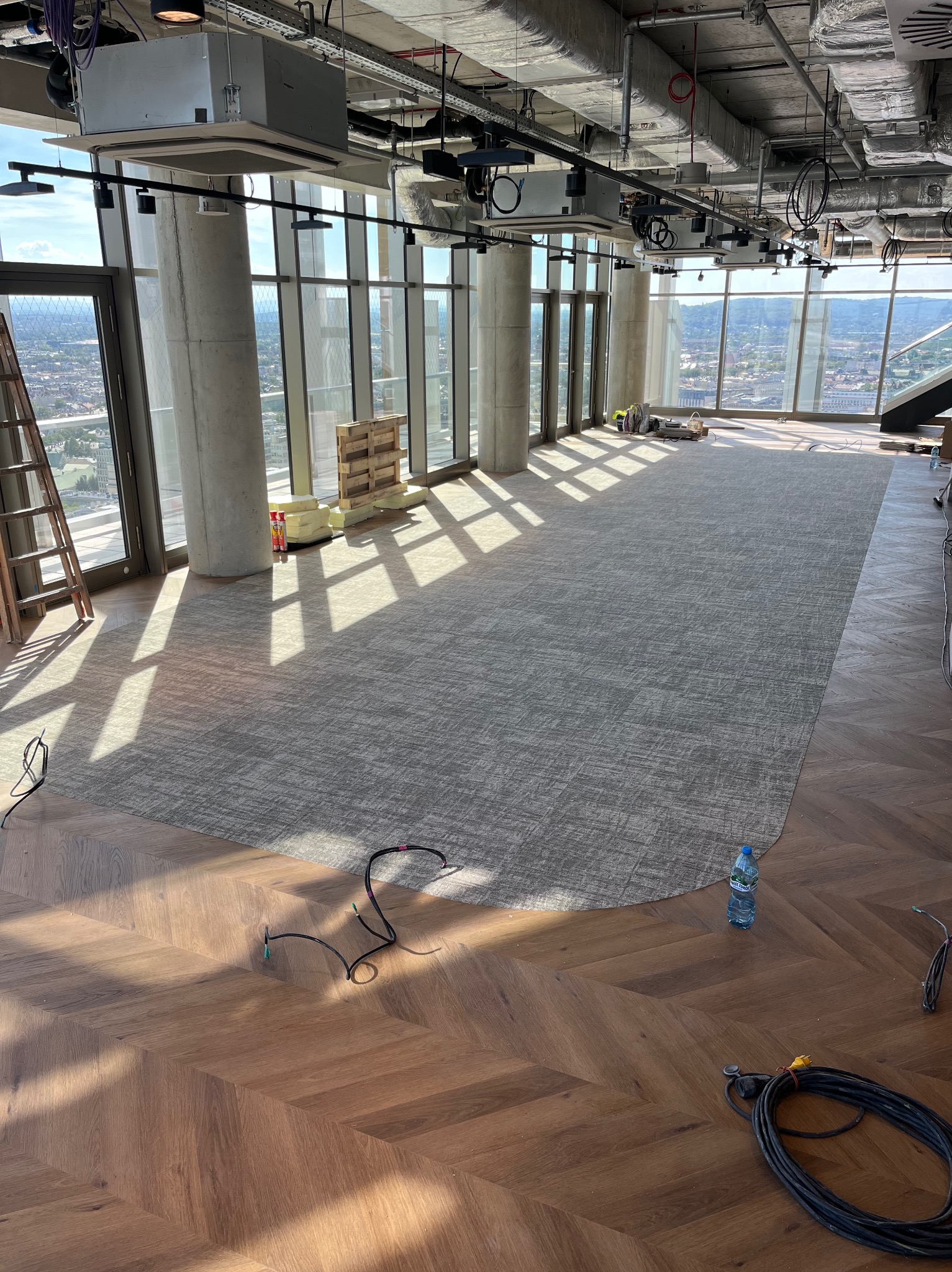 Układanie wykładziny dywanowej w biurze z widokiem na miasto. W tle drabina i materiały budowlane. Na pierwszym planie przewody. Widać światło słoneczne.