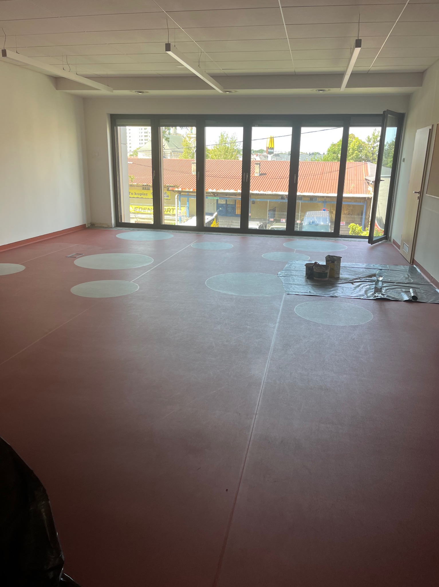 Wnętrze z czerwoną podłogą w białe koła, częściowo zakrytą folią i narzędziami. Duże okna z widokiem na budynki. Prace wykończeniowe.