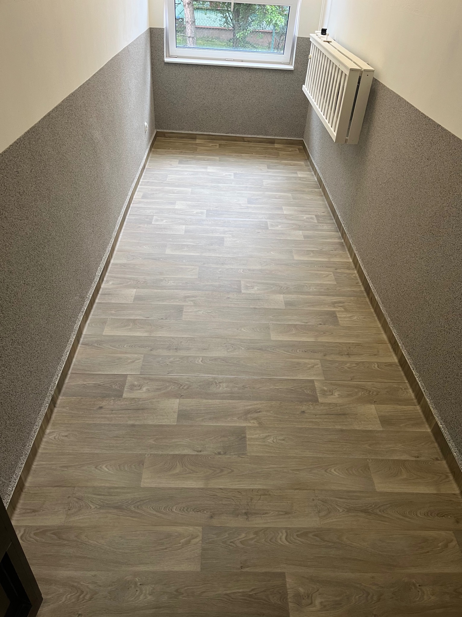 Wąski korytarz z panelami imitującymi drewno na podłodze i szarą, teksturowaną lamperią na ścianach. Widoczne okno i grzejnik składany.