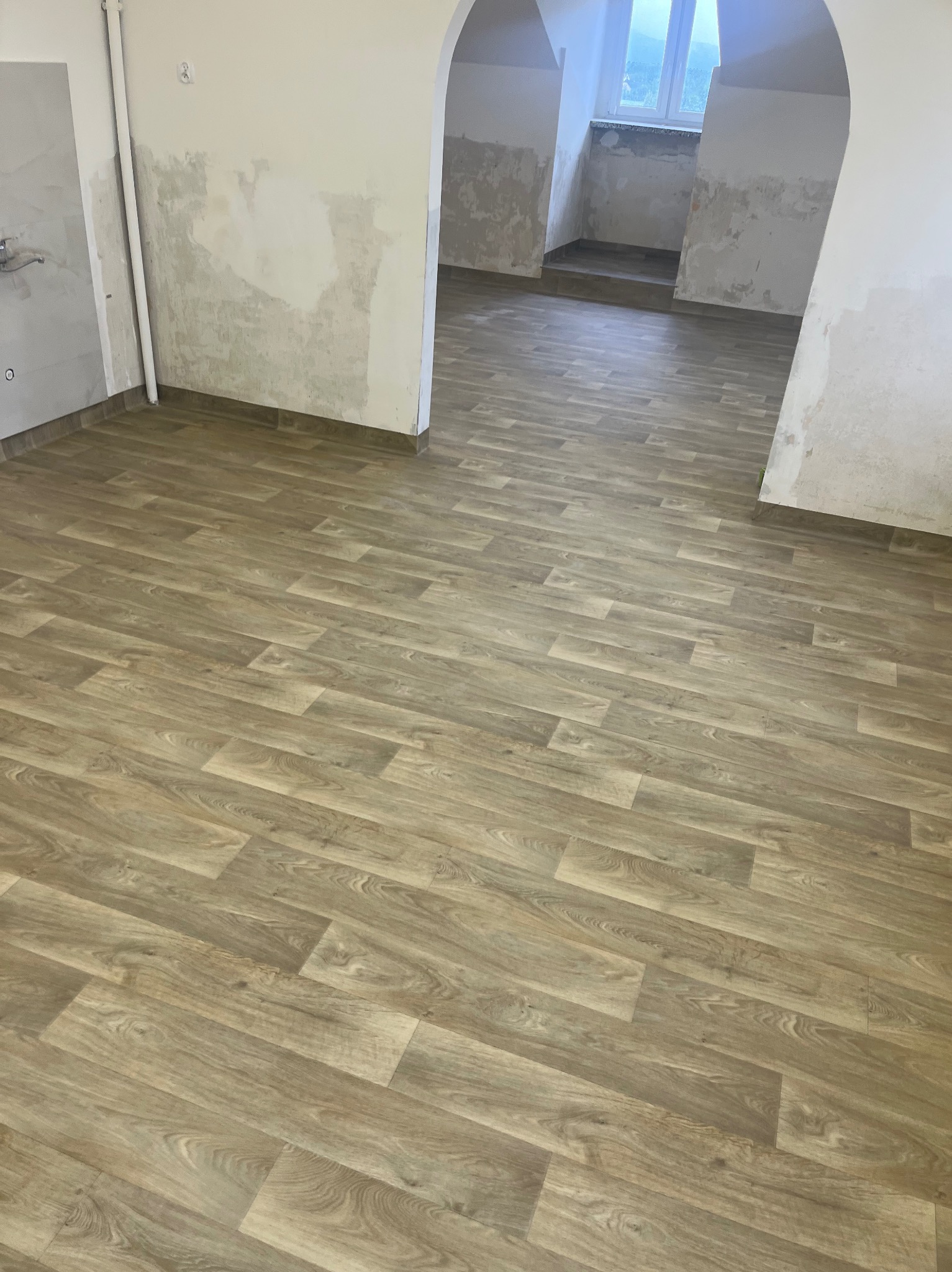 Jasna, drewnopodobna podłoga ułożona w nowym, przestronnym wnętrzu. Ściany częściowo przygotowane do malowania. Widok na kolejne pomieszczenie.