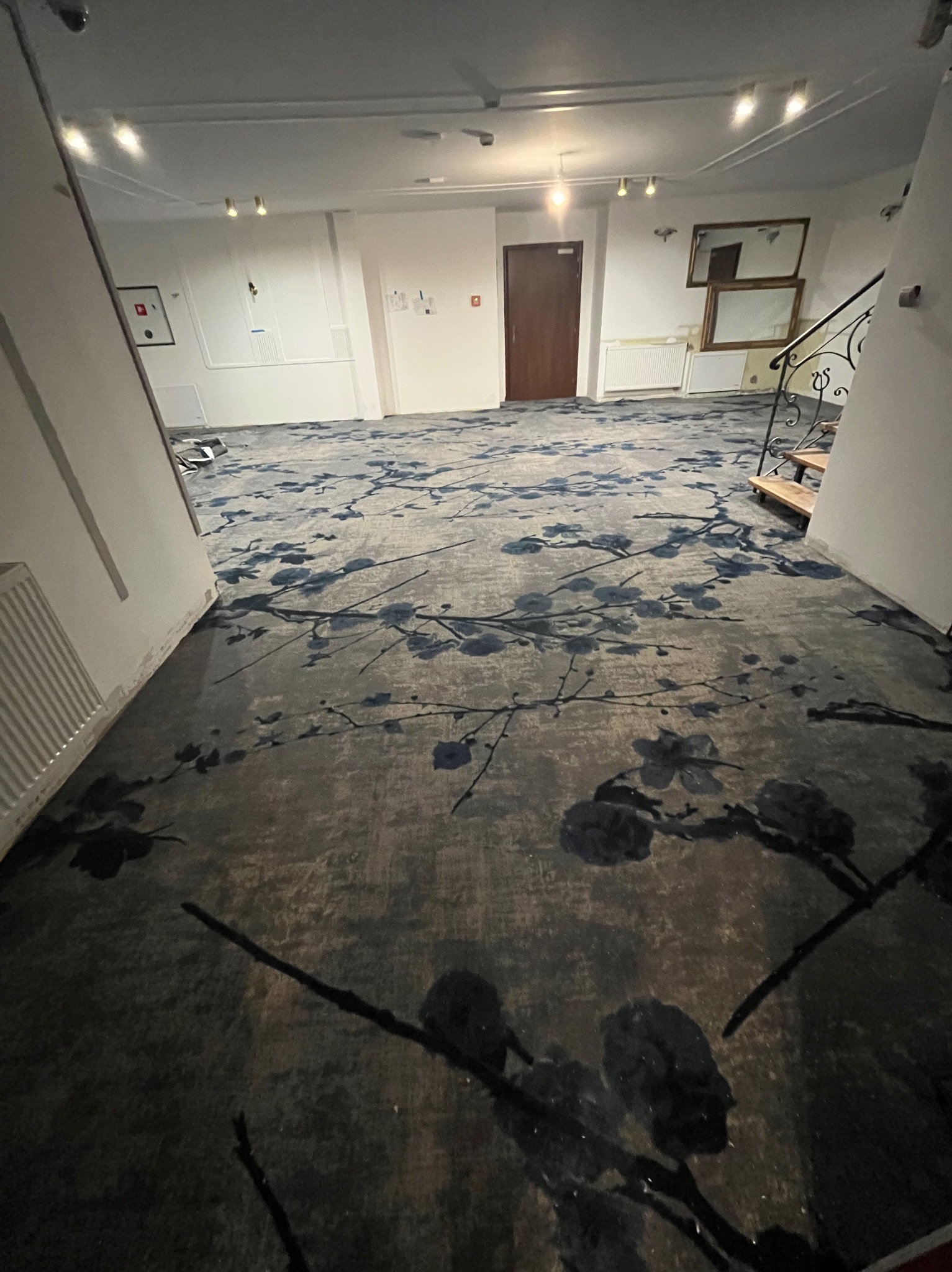 Wnętrze z nową, wzorzystą, szaro-niebieską  podłogą. Widok na korytarz z drzwiami i schodami w tle. Wykładzina w kwiatowy wzór.