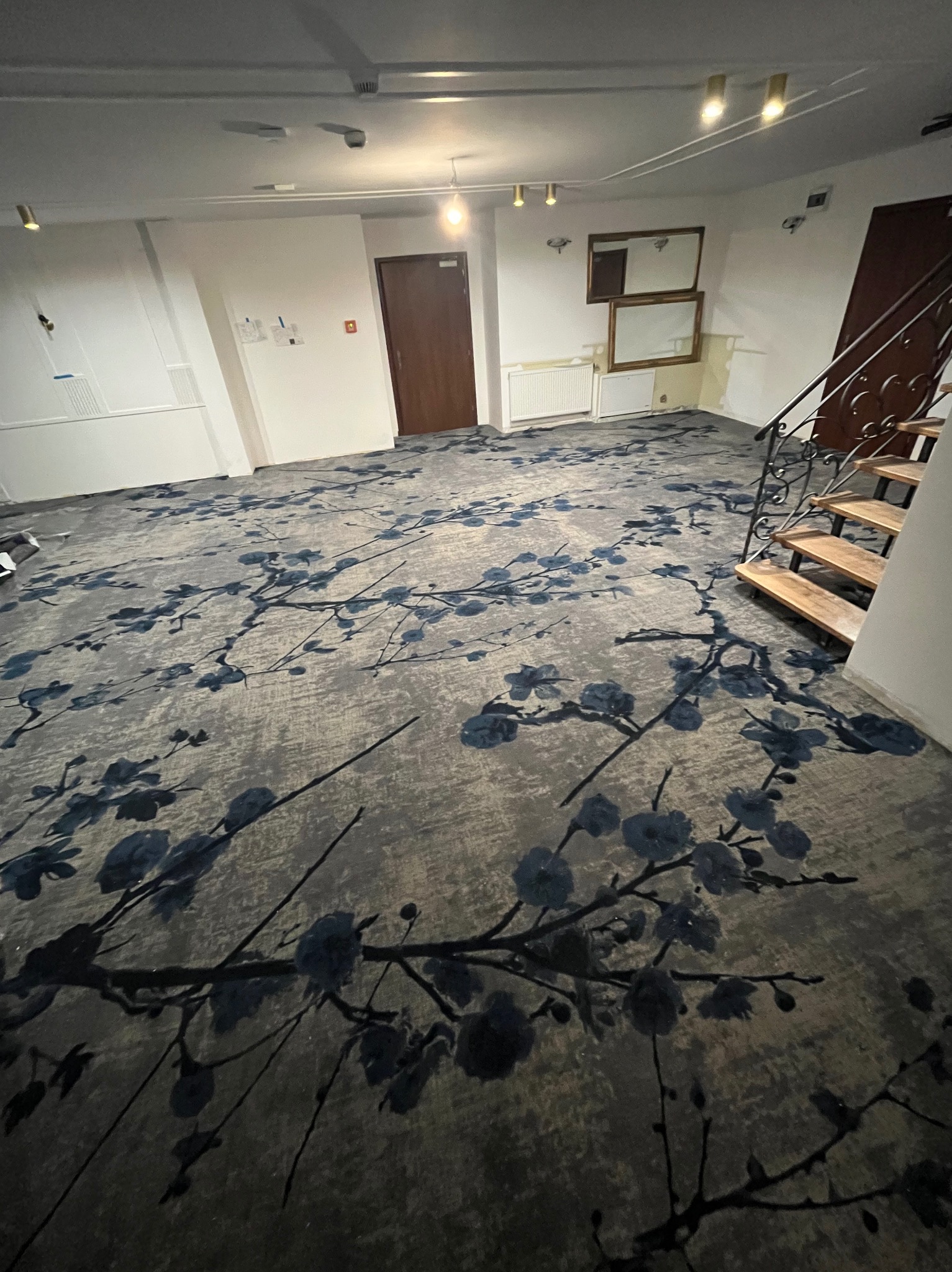 Ukończona podłoga w przestronnym wnętrzu z wykładziną w kwiatowy wzór, widoczne schody z metalową balustradą i drzwi. Nowoczesne oświetlenie sufitowe.