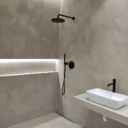 Minimalistyczna łazienka z beżowymi ścianami wykończonymi tynkiem dekoracyjnym, czarną armaturą, deszczownicą i prostokątną umywalką nablatową, oświetlenie LED w niszy.