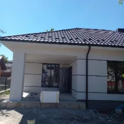 Wykończony dom jednorodzinny z czarnym dachem i jasną elewacją, widoczne materiały budowlane przed wejściem, sugerujące trwające prace wykończeniowe.