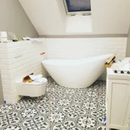 Nowoczesna łazienka na poddaszu w trakcie wykańczania: wolnostojąca wanna, podwieszana toaleta, białe płytki typu metro na ścianach, wzorzysta czarno-biała podłoga, okno dachowe.