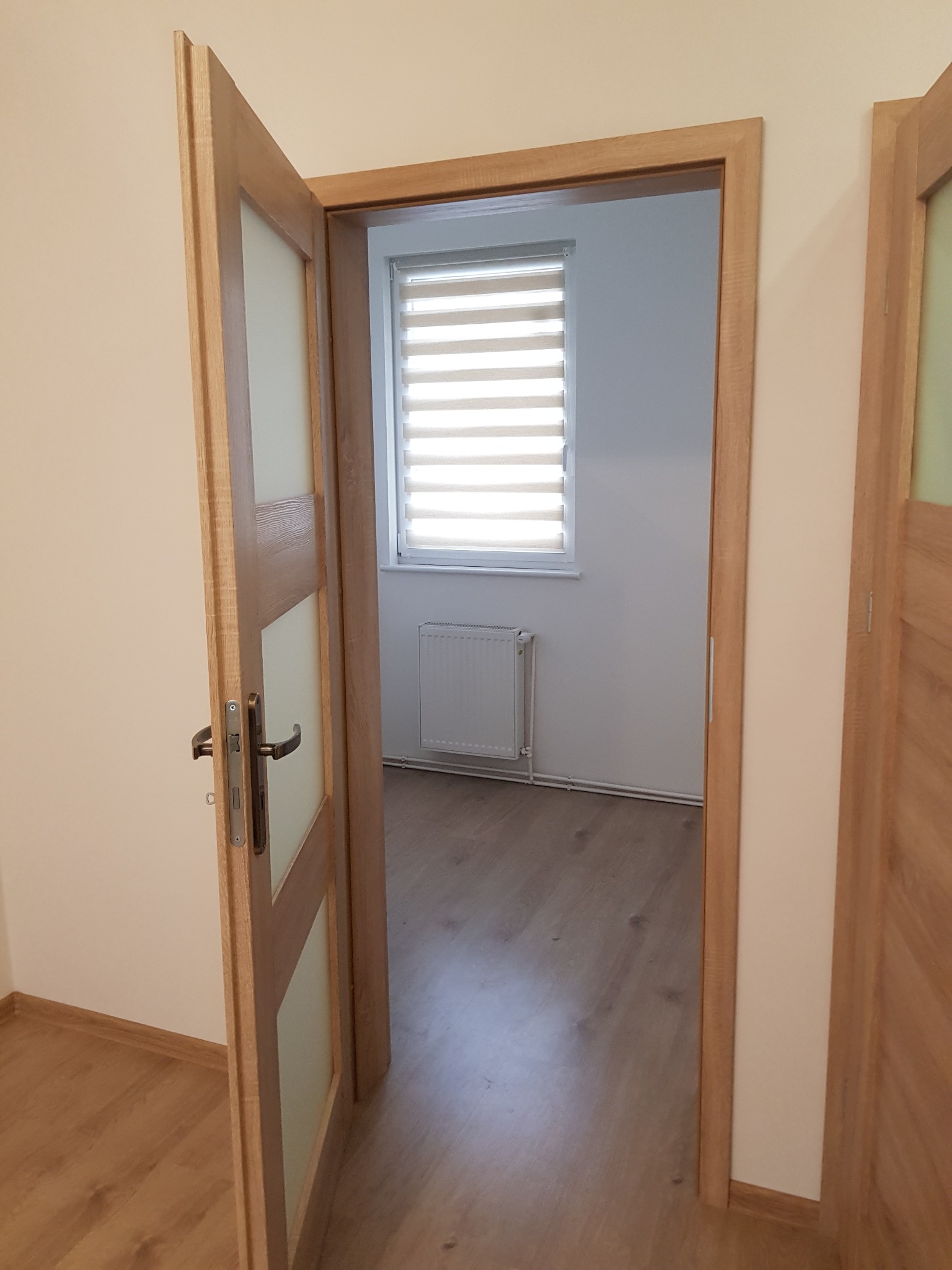 Otwarta drewniana drzwi z przeszkleniami, widok na odnowione wnętrze pokoju z panelami podłogowymi, grzejnikiem i roletami w oknie.