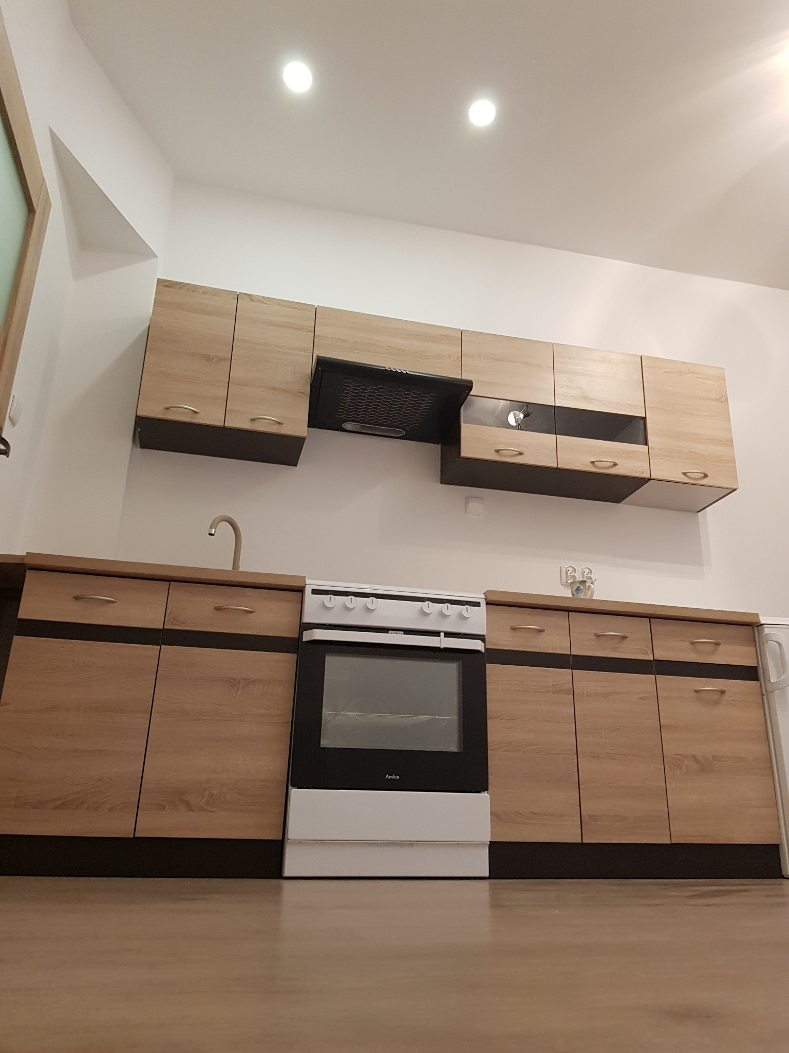 Komplet mebli kuchennych z drewnopodobnymi frontami, czarnymi akcentami i białym sprzętem AGD, oświetlony punktowymi lampami sufitowymi.